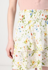 Femme portant une jupe blanche à fleurs vertes, jaunes, rouges et bleues, associée à un haut texturé rose.