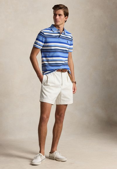 Jeune homme debout les mains dans les poches portant un polo bleu rayé, un short blanc, une ceinture tressée marron et des baskets blanches sur fond neutre.