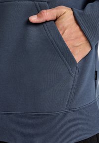 Marineblauwe sweatshirt met een kangoeroezak, ribbels aan de mouwen en de onderrand. Soepele textuur met stikdetails langs de randen.