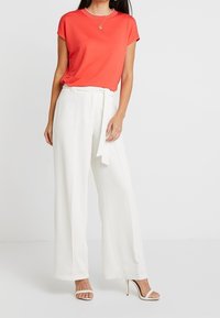 Top rojo de manga corta combinado con pantalones blancos de pierna ancha con cinturón tipo lazo. La modelo lleva tacones blancos con tiras.