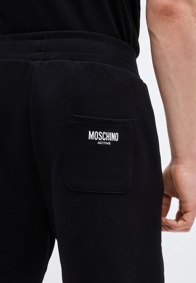 Μαύρα σορτς με πίσω τσέπη και ετικέτα "MOSCHINO ACTIVE", φορεμένα από ένα άτομο με χαλαρό χέρι στο πλάι, μπροστά σε απλό φόντο.