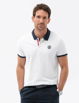 Valkoinen polo-paita, jossa tummansininen ribbikaulus, punaiset ja tummansiniset raidat sekä brodeerattu logo vasemmassa rinnassa. Yhdistetty tummiin housuihin.