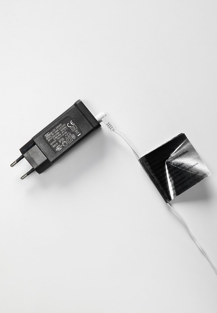 Schwarzes AC-Netzteil mit zwei Stiften und einem weißen Kabel. Ein kleines, teilweise aufgeklapptes schwarzes Heft ist mit dem Kabel verbunden.