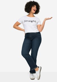 Balta trumpa rankovė t-shirt su juodu tekstu „Straight“, derinama su tamsiai mėlynomis tiesiomis džinsomis ir baltais sportbačiais su pilkais akcentais.