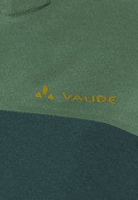 Tessuto verde e scuro con il testo "VAUDE" giallo e logo su materiale per abbigliamento o attrezzatura da esterno.