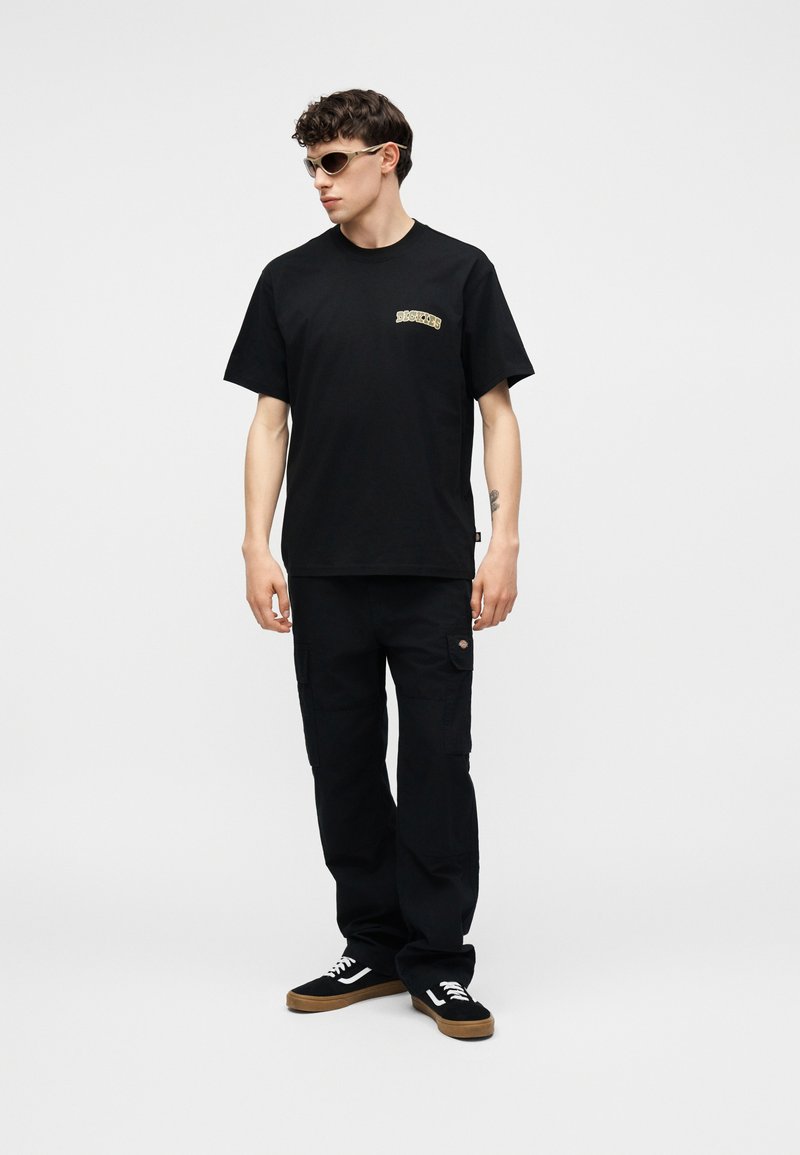 Giovane uomo che indossa una t-shirt nera con grafica, pantaloni cargo neri, sneakers nere e occhiali da sole beige, in piedi su sfondo bianco semplice.