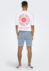 Valkoinen ylisuurikokoinen t-paita, jossa on vaaleanpunainen graafinen teksti "SUNNY VACATION" ja auringon muotoilu selkäpuolella, yhdistettynä vaaleansinisiin farkkushortseihin ja valkoisiin lenkkareihin.