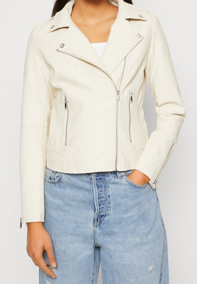 Femme portant une veste en cuir blanche zippée avec des boutons et des fermetures éclair argentés, associée à un jean taille haute bleu clair.