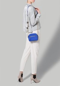 Borsa a tracolla sintetica blu con tasca con zip, tracolla in catena argentata, forma rotonda, abbinata a un top grigio chiaro e pantaloni bianchi.