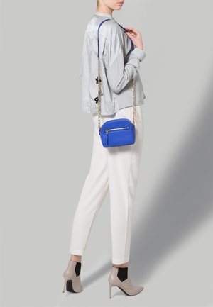 Blå syntetisk crossbody-väska med en dragkedjeficka, silverfärgad kedjesnodd, rundad form, bärs med ljusgrå topp och vita byxor.