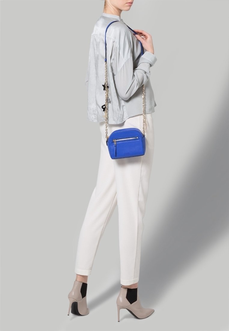 Borsa a tracolla sintetica blu con tasca con zip, tracolla in catena argentata, forma rotonda, abbinata a un top grigio chiaro e pantaloni bianchi.