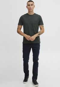 Camiseta de manga corta verde oscura con un bolsillo en el pecho, combinada con pantalones slim-fit azul marino oscuros y zapatillas negras. Diseño sencillo y relajado.