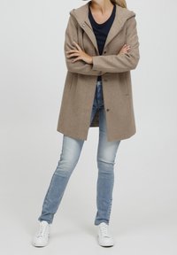 Krémový kabát z vlněné směsi s kapucí, s knoflíkovým zapínáním, bočními kapsami a přímým střihem, spárovaný s světle modrými džíny a bílými teniskami.