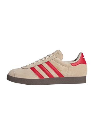 Beige suède sneaker met rode Adidas-strepen, rode hieltab, bruine zool en gouden tekst "Gazelle" aan de zijkant, gezien vanaf de buitenkant.