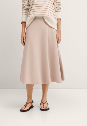 Frau trägt einen beigen A-Linien-Midirock, einen beige-weiß gestreiften Pullover und Sandalen mit geflochtenen Riemen und steht auf einem weißen Boden.