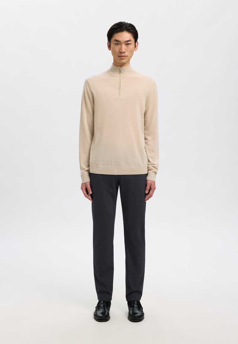 Giovane uomo in piedi rivolto verso avanti indossa un maglione beige a zip, pantaloni neri e scarpe eleganti nere su sfondo bianco.