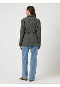 Grijze wollen jas met pied-de-poule patroon en ceintuur in de taille, voorzien van een ritskraag en een op maat gemaakte snit, gecombineerd met blauwe spijkerbroek.