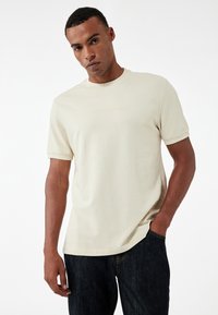 T-shirt beige a maniche corte in tessuto morbido con colletto a costine e testo in rilievo sul petto. Abbinato a jeans scuri.