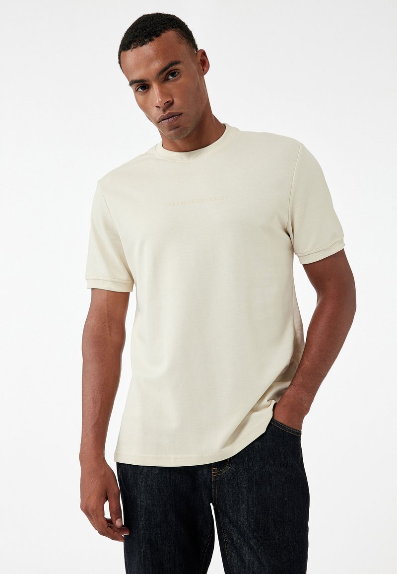 T-shirt beige a maniche corte in tessuto morbido con colletto a costine e testo in rilievo sul petto. Abbinato a jeans scuri.