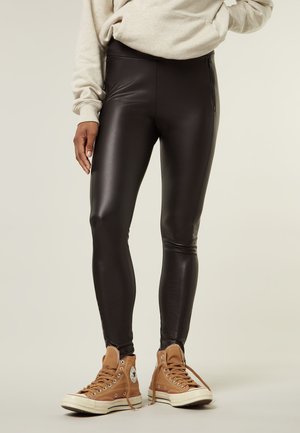 Leggings in finta pelle nera con vita alta e tasche laterali con zip, abbinati a sneakers alte beige. Texture liscia con un design aderente.