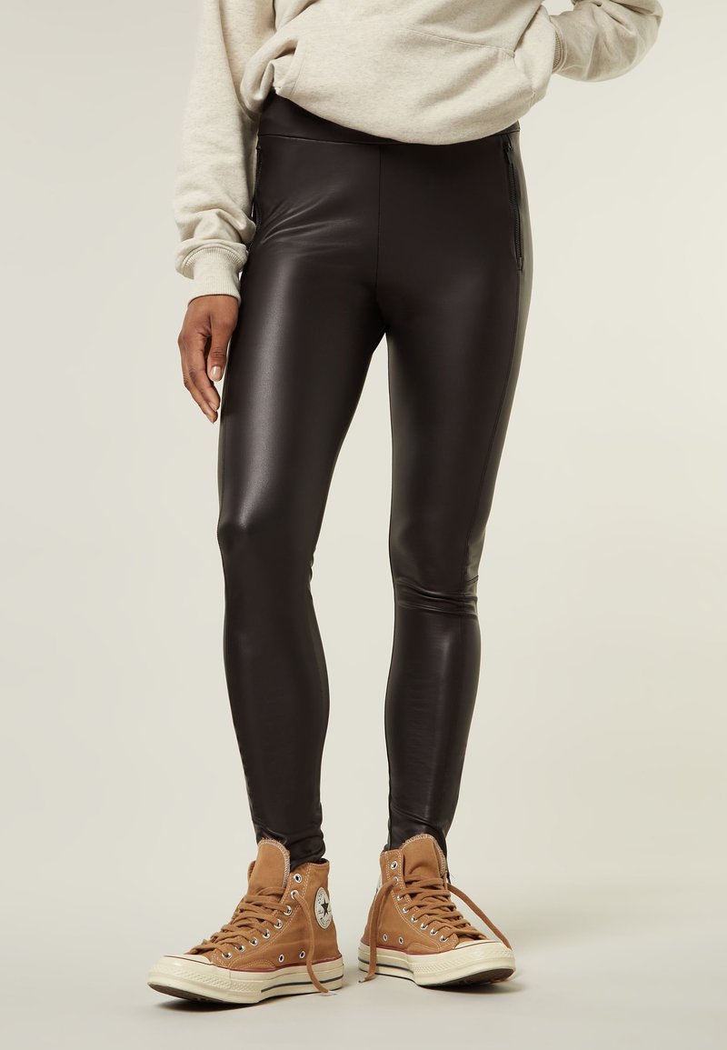 Leggings in finta pelle nera con vita alta e tasche laterali con zip, abbinati a sneakers alte beige. Texture liscia con un design aderente.