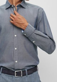 Camicia con bottoni in tessuto chambray blu, con colletto a punta e bottoni bianchi. Le maniche lunghe presentano polsini con bottoni.