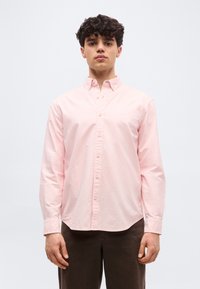 Jeune homme aux cheveux bouclés portant une chemise rose clair à boutons et un pantalon marron foncé, debout devant un fond blanc uni.