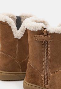 Stivaletti alla caviglia in suede marrone con fodera in pelliccia bianca nella parte superiore, dotati di una cerniera laterale e dettagli di cucitura lungo le cuciture. Suola in gomma piatta.