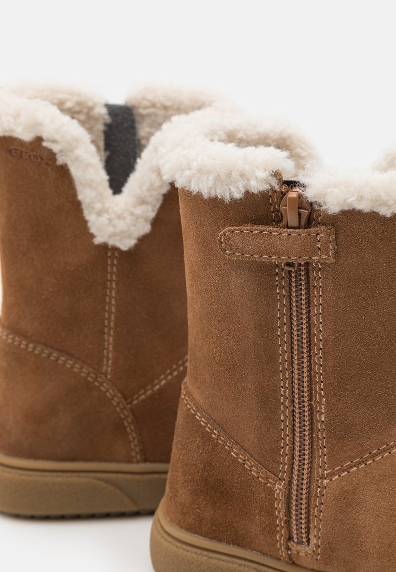 Braune Wildleder-Ankle-Boots mit weißem Fellfutter oben, ausgestattet mit einem seitlichen Reißverschluss und Nähdetails entlang der Nähte. Flacher Gummisohlen. 