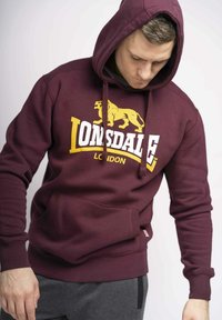 Felpa bordeaux con tasca frontale, logo Lonsdale e grafica di un leone in giallo. Presenta un cappuccio con lacci e polsini a costine. Realizzata in morbido misto cotone.