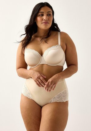 Kurvet kvinde iført beige bh med blonder og højhættede trusser, stående med hænderne let foldet, og kigger svagt til siden.