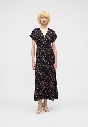 Femme aux cheveux blonds courts et bouclés, portant une robe noire longue jusqu'aux chevilles avec de petites fleurs roses imprimées, et des sandales à talons beiges, debout de face.