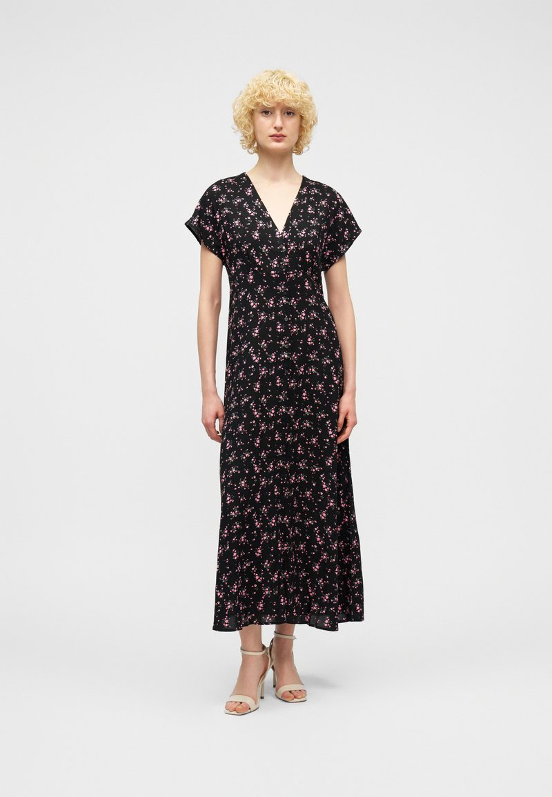 Γυναίκα με κοντά κατσαρά ξανθά μαλλιά, φορώντας μαύρο φόρεμα μέχρι τον αστράγαλο με μικρά ροζ ανθισμένα σχέδια και μπεζ σανδάλια με τακούνι, στέκεται με το σώμα προς τα εμπρός.