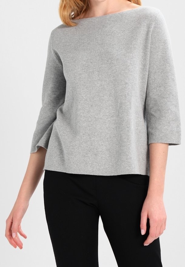 Femme portant un pull gris clair à manches longues et à encolure bateau et un pantalon noir, debout devant un fond clair uni.