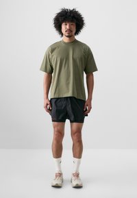 Olivově zelené bavlněné tričko s volným střihem, černé šortky s červenými akcenty a světlé sportovní boty. Minimalistický design, standardní délka.
