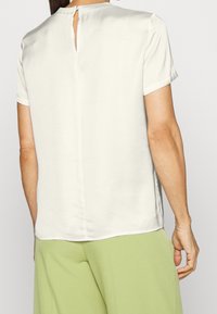 Haut beige clair à manches courtes, légèrement ample, avec une ouverture en forme de clef dans le dos et une texture lisse et soyeuse.