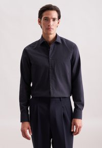 Camicia a maniche lunghe blu scuro realizzata in un tessuto di struttura, con colletto button-down e taschino sul petto. Indossata con pantaloni pieghettati scuri.