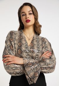 Slangeprint top med V-hals, lange bløde ærmer og perler. Farverne inkluderer beige, brun og sort. Modellen står med armene over kors.