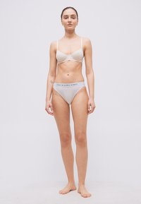 Γυναίκα που στέκεται ξυπόλυτη, φοράει ανοιχτό μπεζ σουτιέν και λευκά εσώρουχα Polo Ralph Lauren, με φόντο απλό λευκό.