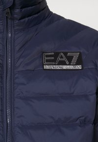 Marinblå puffer-jacka med dragkedja, kviltad textur och en framträdande svart logopatch med "EA7 Emporio Armani"-märkning.