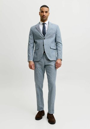 Homme portant un costume ajusté gris clair, une chemise blanche, une cravate sombre à motifs et des mocassins marron, debout devant un fond clair uni.