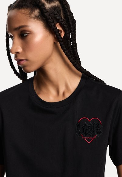 Giovane con capelli intrecciati che indossa una maglietta nera con un cuore rosso e la parola "LOVE" ricamata sul petto.
