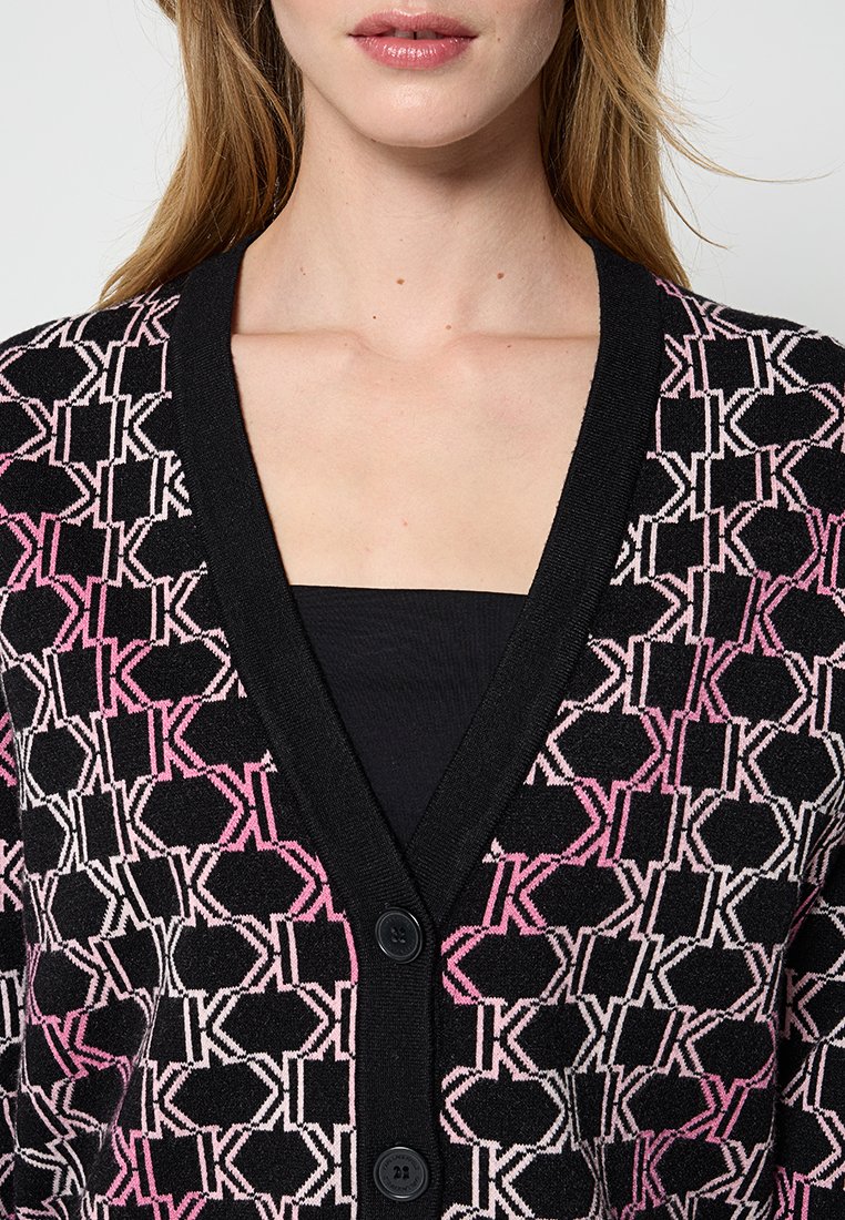Strickjacke mit schwarzem Grundton und einem sich wiederholenden geometrischen Muster in Pink und Weiß, V-Ausschnitt und zwei schwarzen Knöpfen.