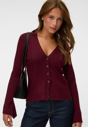 Femme aux longs cheveux bruns portant des boucles d’oreilles en perles, un cardigan bordeaux boutonné, un jean foncé et un sac à bandoulière noir.
