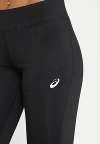 Zbliżenie osoby w czarnych legginsach sportowych z białym logo ASICS na górnej części uda na białym tle.