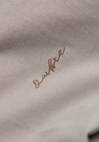 Světlý béžový materiál se vyšívaným kurzívním textem v tmavším béžovém odstínu. Hladká textura s jemným tkaným vzorem.