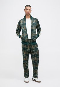 Trening verde realizat dintr-un material moale, cu un model ornate de culoare aurie. Include o jachetă cu fermoar și pantaloni asortați cu dungi albe.