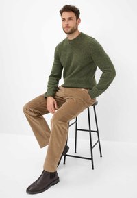 Maglione a maglia verde maculato, pantaloni in corduroy beige e stivali alla caviglia in pelle marrone, seduto su uno sgabello in metallo nero su uno sfondo bianco.