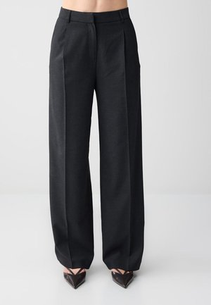 Pantalon noir taille haute à jambes larges avec plis avant, porté avec des chaussures noires à bout pointu sur un fond neutre.