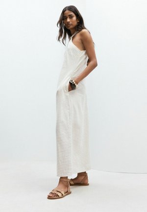 Woman standing wearing a sleeveless, light-striped maxi dress with pockets, sandals, and beaded bracelets against a plain white background.